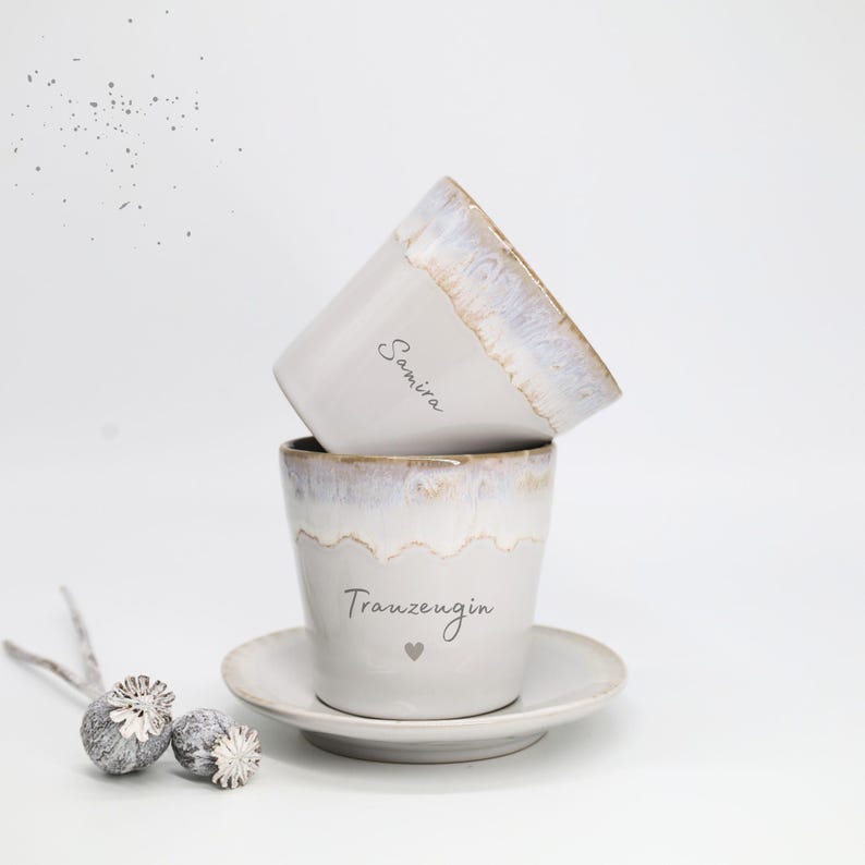 May include: Two white ceramic cups with a light brown rim and the names "Samira" and "Trauzeugin" printed on them. One cup is stacked on top of the other. A small saucer is under the bottom cup. A decorative element is on the left.