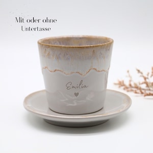 May include: A ceramic coffee cup and saucer set. The cup is white with a light brown rim and the name "Emilia" and a heart etched on the side. The saucer is a matching light gray color. Text above the cup reads "Mit oder ohne Untertasse".