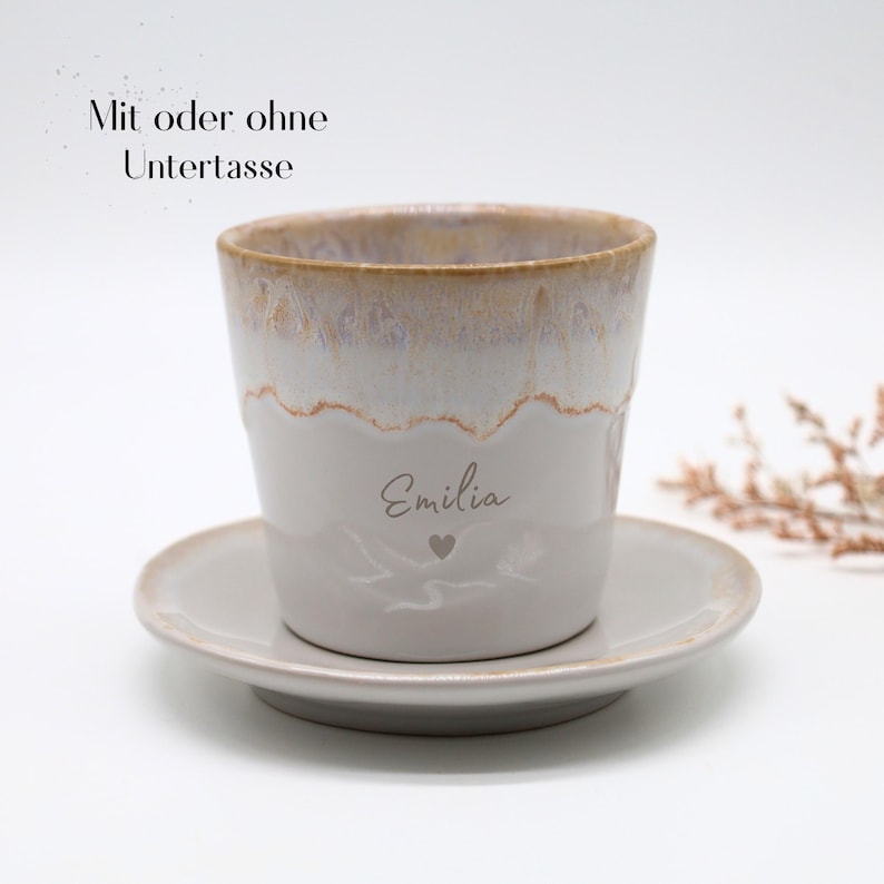 May include: A ceramic coffee cup and saucer set. The cup is white with a light brown rim and the name "Emilia" and a heart etched on the side. The saucer is a matching light gray color. Text above the cup reads "Mit oder ohne Untertasse".