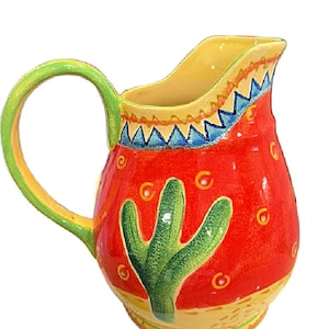 May include: A hand-painted ceramic pitcher with a vibrant Southwestern design. The pitcher features a red body with a green cactus, yellow and blue accents, and a green handle. The pitcher is approximately 8 inches tall.