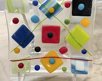 May include: A clear glass decorative plate with colorful geometric shapes. The plate features squares, diamonds, and circles in various colors, including blue, red, yellow, green, and black. The design is abstract and modern, with a clear glass stand.