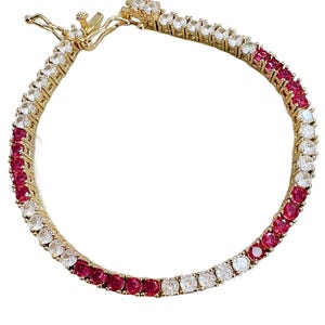 May include: A gold-toned tennis bracelet with alternating white and red gemstones. The gemstones are round-cut and set closely together, creating a continuous line of sparkle. The clasp is also gold-toned and features additional gemstones.