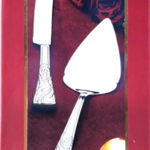 May include: A Lenox holiday cake knife and server set. The set includes a cake knife and server with ornate silver handles and white blades. The background features a burgundy color with decorative elements and the text "HOLIDAY CAKE KNIFE AND SERVER".
