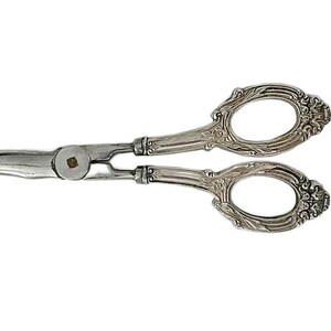 May include: Silver-toned metal scissors featuring curved blades and ornate, embossed handles with intricate floral and leaf detailing. The central pivot point is a circular metal fastener, and the finger loops are oval-shaped with decorative edges.
