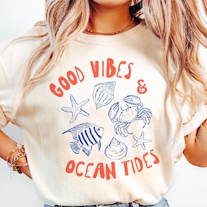 Pu&ograve; includere: Una t-shirt color crema con una stampa grafica che riporta il testo "GOOD VIBES & OCEAN TIDES" in rosso. Il design include illustrazioni a linee blu di una stella marina, una conchiglia, un granchio, un pesce a strisce e una piccola stella.