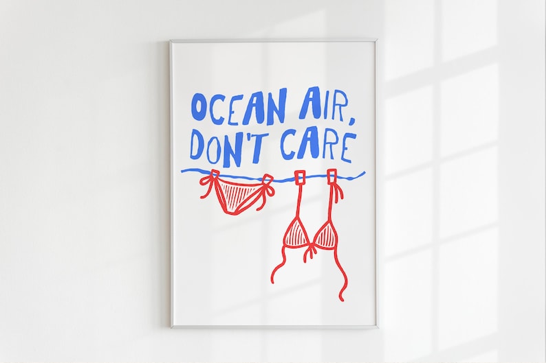 Pu&ograve; includere: Una stampa artistica incorniciata con il testo blu "OCEAN AIR, DON'T CARE" sopra una linea orizzontale blu. Due pezzi di bikini rossi sono appesi alla linea con delle mollette, raffigurati in un semplice stile a linee su sfondo bianco.