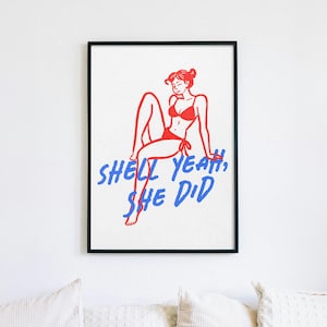 Pu&ograve; includere: Una stampa artistica incorniciata che presenta un disegno a linee rosse di una persona in bikini seduta, accompagnata dal testo blu SHELL YEAH, SHE DID. L'opera &egrave; presentata in una sottile cornice nera su uno sfondo bianco.