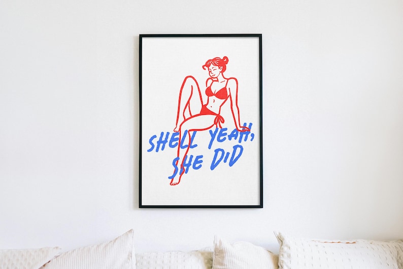 Pu&ograve; includere: Una stampa artistica incorniciata che presenta un disegno a linee rosse di una persona in bikini seduta, accompagnata dal testo blu SHELL YEAH, SHE DID. L'opera &egrave; presentata in una sottile cornice nera su uno sfondo bianco.