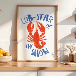 Pu&ograve; includere: Una stampa artistica da parete rettangolare in una cornice di legno chiaro con un'illustrazione di un'aragosta arancione brillante centrata su sfondo bianco con il testo blu LOB-STAR OF THE SHOW arcuato sopra e sotto in stile disegnato.
