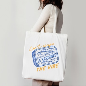 Pu&ograve; includere: Una borsa tote in tela bianca con una grafica blu di una scatola di sardine, con il testo giallo "Can't escape THE VIBE" e le scritte blu "SALT WATER LE SARDINES SARDINIA" e "FRESH FROM PORTUGAL" stampate sul davanti.