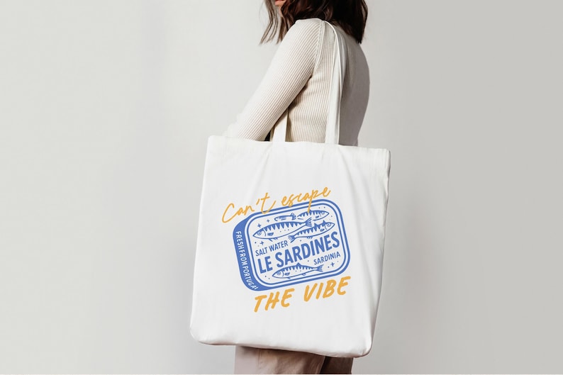Pu&ograve; includere: Una borsa tote in tela bianca con una grafica blu di una scatola di sardine, con il testo giallo "Can't escape THE VIBE" e le scritte blu "SALT WATER LE SARDINES SARDINIA" e "FRESH FROM PORTUGAL" stampate sul davanti.