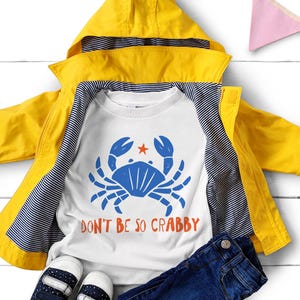 Pu&ograve; includere: Una giacca con cappuccio giallo brillante sopra una t-shirt bianca con un grafico di un granchio blu, una piccola stella arancione e il testo "DON'T BE SO CRABBY" in arancione. La giacca ha una fodera a righe blu navy e bianche, con jeans blu scuro.