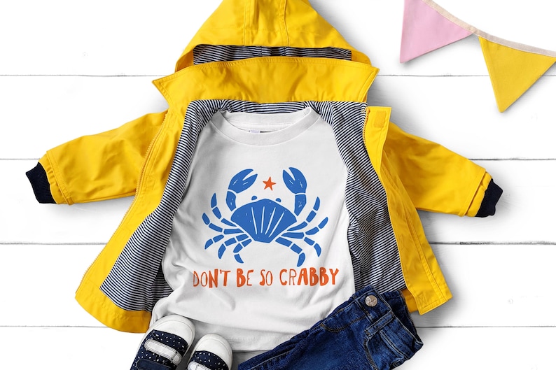 Pu&ograve; includere: Una giacca con cappuccio giallo brillante sopra una t-shirt bianca con un grafico di un granchio blu, una piccola stella arancione e il testo "DON'T BE SO CRABBY" in arancione. La giacca ha una fodera a righe blu navy e bianche, con jeans blu scuro.