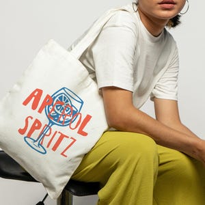 Pu&ograve; includere: Una borsa tote in tela bianca con una grafica blu di un bicchiere da cocktail con una fetta di agrume e il testo rosso "APEROL SPRITZ" stampato sul davanti. La borsa ha una forma rettangolare semplice con una tracolla e fondo piatto.
