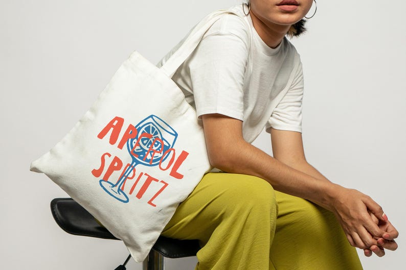 Pu&ograve; includere: Una borsa tote in tela bianca con una grafica blu di un bicchiere da cocktail con una fetta di agrume e il testo rosso "APEROL SPRITZ" stampato sul davanti. La borsa ha una forma rettangolare semplice con una tracolla e fondo piatto.