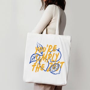 Pu&ograve; includere: Una borsa tote in tela bianca con un design grafico che presenta contorni blu di limoni e foglie. Il testo "YOU'RE SIMPLY THE ZEST" &egrave; stampato in un carattere giallo audace al centro della borsa, integrato con le illustrazioni dei limoni.