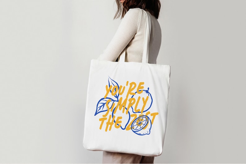 Pu&ograve; includere: Una borsa tote in tela bianca con un design grafico che presenta contorni blu di limoni e foglie. Il testo "YOU'RE SIMPLY THE ZEST" &egrave; stampato in un carattere giallo audace al centro della borsa, integrato con le illustrazioni dei limoni.