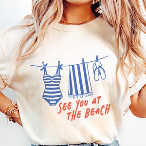 Pu&ograve; includere: Una t-shirt a maniche corte color crema con una grafica blu che raffigura un costume da bagno a righe, un asciugamano a righe e infradito appesi a un filo da bucato. Sotto la grafica, il testo "SEE YOU AT THE BEACH" &egrave; scritto in rosso.