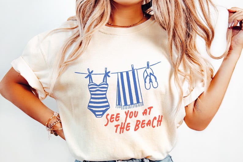 Pu&ograve; includere: Una t-shirt a maniche corte color crema con una grafica blu che raffigura un costume da bagno a righe, un asciugamano a righe e infradito appesi a un filo da bucato. Sotto la grafica, il testo "SEE YOU AT THE BEACH" &egrave; scritto in rosso.