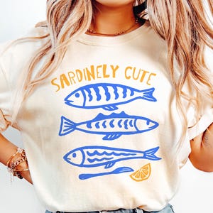 Pu&ograve; includere: Una t-shirt a maniche corte color crema con una stampa grafica di tre pesci blu impilati verticalmente. Sopra i pesci, il testo SARDINELY CUTE &egrave; stampato in giallo. Sotto i pesci, ci sono uno spicchio di limone giallo e un utensile blu.