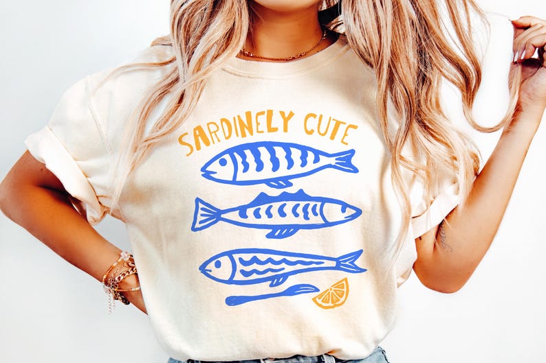Pu&ograve; includere: Una t-shirt a maniche corte color crema con una stampa grafica di tre pesci blu impilati verticalmente. Sopra i pesci, il testo SARDINELY CUTE &egrave; stampato in giallo. Sotto i pesci, ci sono uno spicchio di limone giallo e un utensile blu.