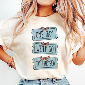 May include: Cream-colored t-shirt with the text "ONE DAY WE'LL GO TO THE SEA" in a blue ticket design. Each ticket has a pink bow. The shirt is a casual, everyday wear item.