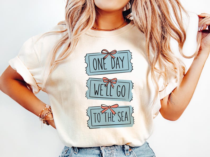 May include: Cream-colored t-shirt with the text "ONE DAY WE'LL GO TO THE SEA" in a blue ticket design. Each ticket has a pink bow. The shirt is a casual, everyday wear item.