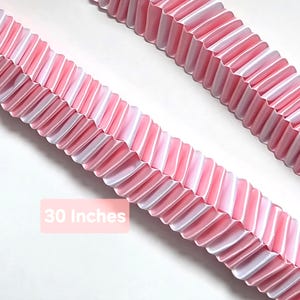 May include: Two strips of pink and white pleated paper garland, each measuring 30 inches in length. The paper features a repeating accordion-fold pattern with alternating pink and white vertical stripes, creating a textured, decorative linear design.