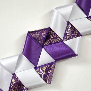 May include: A decorative ribbon braid featuring a repeating geometric pattern. The design consists of alternating segments of solid white satin ribbon, solid purple satin ribbon, and purple satin ribbon with a metallic gold filigree scroll print.