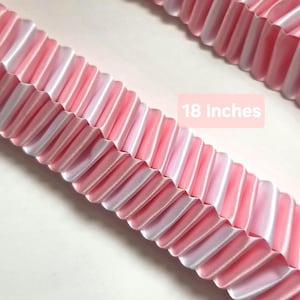 May include: A pleated ribbon in a light pink shade with a smooth, satin-like finish. The ribbon features a repeating vertical fold pattern throughout its length. A pink rectangular label with the white text 18 Inches is visible.