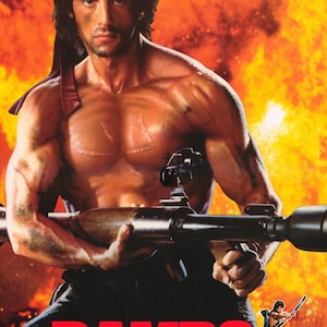 May include: Movie poster for "Rambo: First Blood Part II" featuring a muscular man holding a weapon against a fiery background. The title "RAMBO" is in bold red letters, with the tagline "FIRST BLOOD PART II" below.