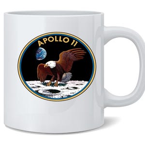 May include: White ceramic mug featuring the Apollo 11 mission emblem. The design includes a brown and white eagle on the moon, with the Earth in the background, and the words "Apollo 11" in gold lettering.