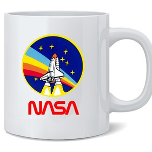 May include: White ceramic mug featuring the NASA logo. The logo includes a space shuttle graphic with a rainbow arc and stars, set against a blue background. The word "NASA" is printed in red below the logo. The mug has a curved handle.