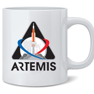 May include: White ceramic mug featuring the Artemis program logo. The logo includes a rocket launching from a moon graphic, with a black triangle and the word "ARTEMIS" in bold black letters. The mug has a curved handle.