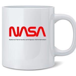 May include: White ceramic mug featuring the NASA logo in red, with the words "National Aeronautics and Space Administration" printed below. The mug has a curved handle and a glossy finish.