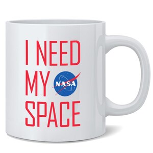 May include: A white ceramic mug featuring the red text I NEED MY SPACE printed vertically on the front. The word SPACE includes the blue and white circular NASA logo as the letter A. The mug has a standard handle on the right side.