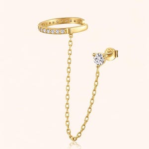 Può includere: Un set di orecchini e polsino per l'orecchio color oro. Il polsino per l'orecchio presenta una fila di piccole pietre trasparenti. Una delicata catena collega il polsino per l'orecchio a un orecchino con una singola pietra rotonda e trasparente.