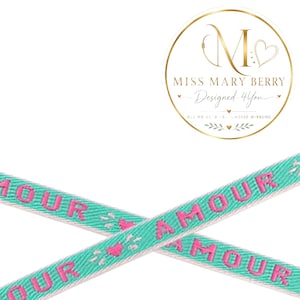 Könnte beinhalten: Zwei gekreuzte türkisfarbene Bänder mit dem in rosa Garn gestickten Wort AMOUR. Kleine rosa Herzsymbole sind zwischen dem Text platziert. Ein kreisförmiges Logo mit dem Text MISS MARY BERRY Designed 4You ist ebenfalls zu sehen.
