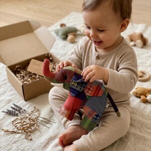 Op de afbeelding: Een kleurrijk patchwork olifantenspeelgoed met een rode slurf en witte slagtanden. Het speelgoed wordt vastgehouden door een baby in een beige onesie. Een kartonnen doos en ander houten speelgoed staan op de achtergrond.
