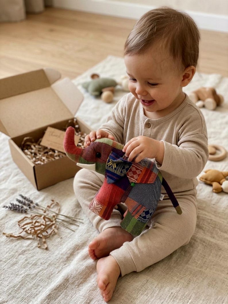 Op de afbeelding: Een kleurrijk patchwork olifantenspeelgoed met een rode slurf en witte slagtanden. Het speelgoed wordt vastgehouden door een baby in een beige onesie. Een kartonnen doos en ander houten speelgoed staan op de achtergrond.