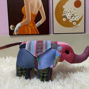Op de afbeelding: Een patchwork olifant knuffel met een roze kop en slurf, een blauwe spijkerbroek body en patroonstoffen accenten in rood, blauw en groen. Het speelgoed heeft een klein groen belletje en witte slagtanden. Het wordt gepresenteerd op een witte, zachte ondergrond.