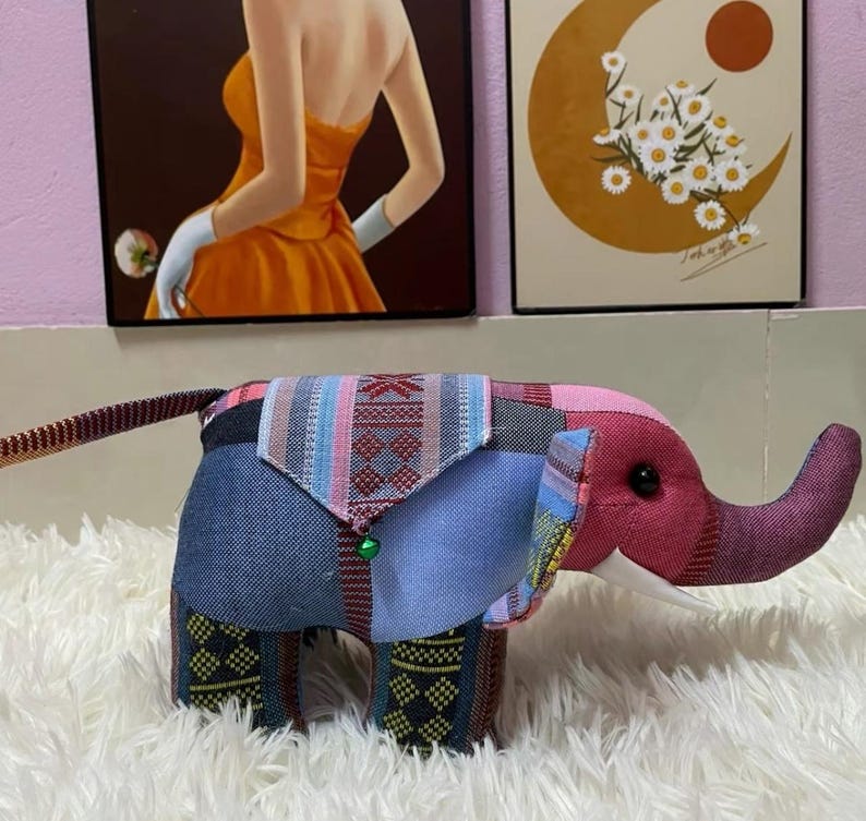 Op de afbeelding: Een patchwork olifant knuffel met een roze kop en slurf, een blauwe spijkerbroek body en patroonstoffen accenten in rood, blauw en groen. Het speelgoed heeft een klein groen belletje en witte slagtanden. Het wordt gepresenteerd op een witte, zachte ondergrond.