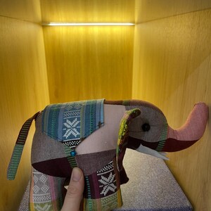 Op de afbeelding: Een handgemaakte olifant knuffel met een patchwork ontwerp. De olifant heeft een roze slurf, oren en lichaam, met stof met patronen in blauwe, groene en bordeaux tinten. Het speelgoed heeft kleine witte slagtanden en een kralen accent.