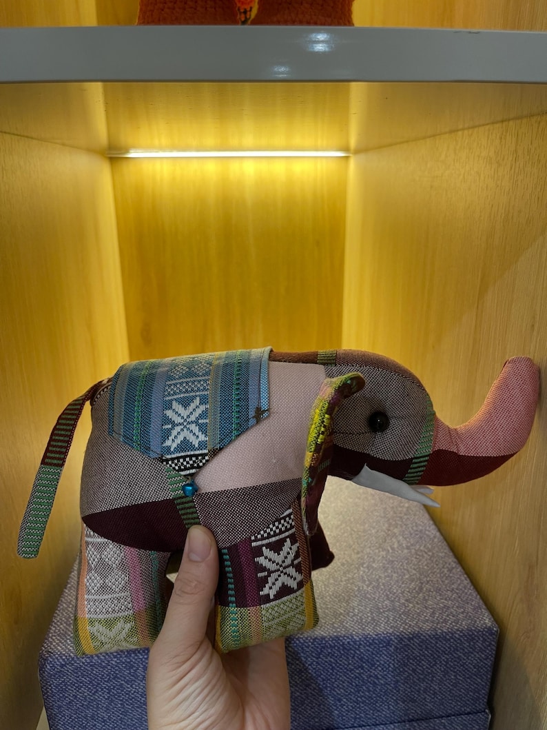 Op de afbeelding: Een handgemaakte olifant knuffel met een patchwork ontwerp. De olifant heeft een roze slurf, oren en lichaam, met stof met patronen in blauwe, groene en bordeaux tinten. Het speelgoed heeft kleine witte slagtanden en een kralen accent.