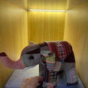 Op de afbeelding: Een patchwork olifant speelgoed in tinten bruin, rood en roze. De olifant heeft een lange slurf en grote oren. Het speelgoed staat op een blauw oppervlak.