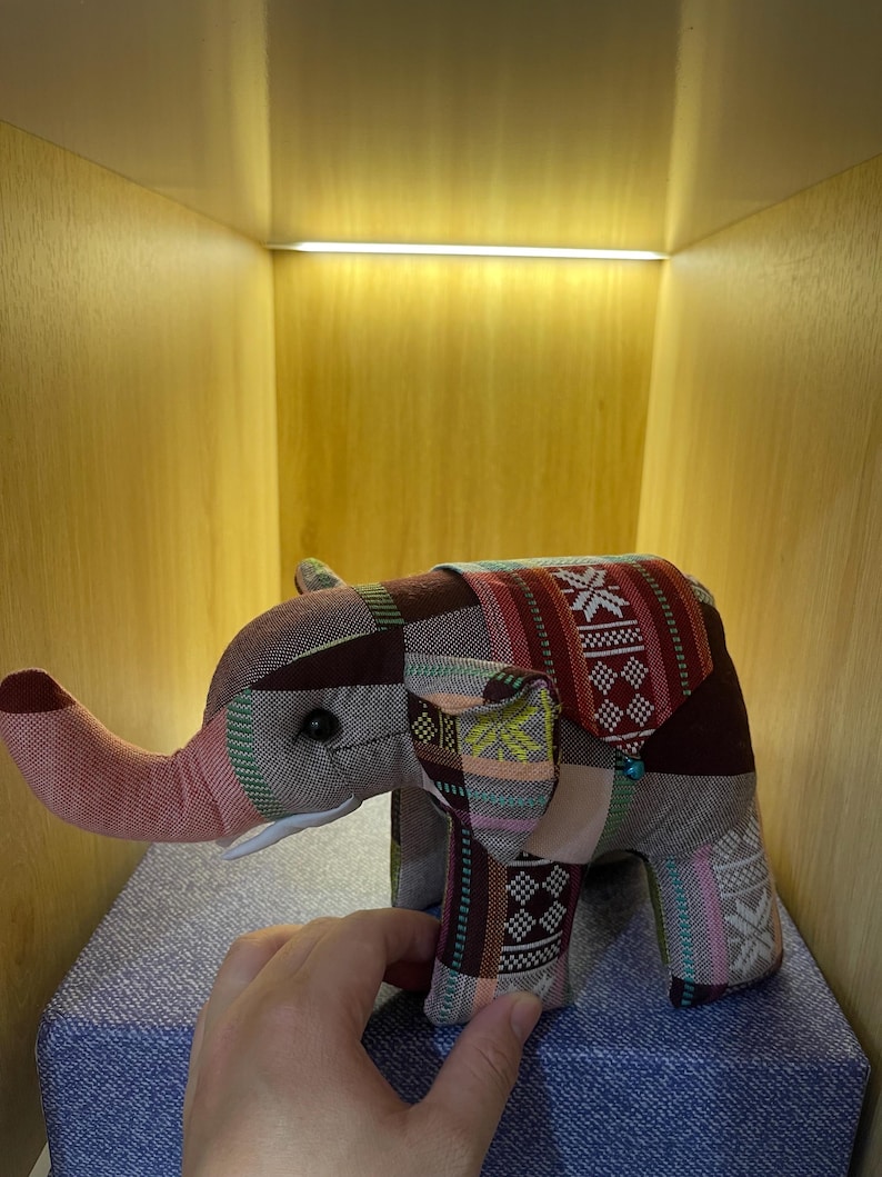Op de afbeelding: Een patchwork olifant speelgoed in tinten bruin, rood en roze. De olifant heeft een lange slurf en grote oren. Het speelgoed staat op een blauw oppervlak.