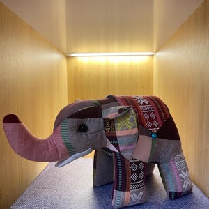 Op de afbeelding: Een stoffen olifant speelgoed met een patchwork ontwerp in tinten bordeaux, roze en groen. De olifant heeft een lange slurf, kleine zwarte ogen en een turquoise knoop. Het speelgoed staat op een blauw oppervlak.
