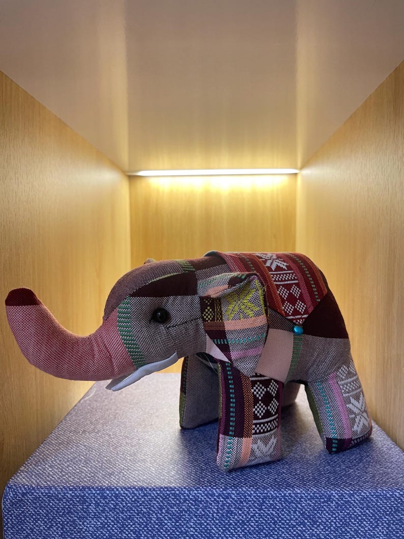 Op de afbeelding: Een stoffen olifant speelgoed met een patchwork ontwerp in tinten bordeaux, roze en groen. De olifant heeft een lange slurf, kleine zwarte ogen en een turquoise knoop. Het speelgoed staat op een blauw oppervlak.
