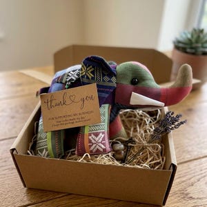 Op de afbeelding: Een handgemaakte stoffen olifant met een kleurrijk patchwork-ontwerp, gepresenteerd in een bruine kartonnen doos gevuld met stro. Een klein kaartje met "dank je" en een takje lavendel zijn inbegrepen.