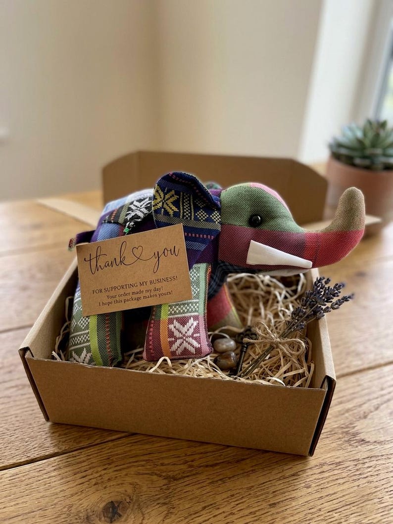 Op de afbeelding: Een handgemaakte stoffen olifant met een kleurrijk patchwork-ontwerp, gepresenteerd in een bruine kartonnen doos gevuld met stro. Een klein kaartje met "dank je" en een takje lavendel zijn inbegrepen.